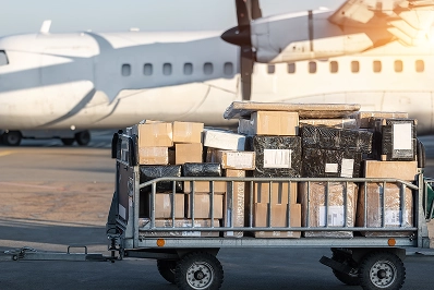 Wózek bagażowy z różnymi pudełkami i paczkami przed stojącym samolotem na lotnisku w ciepłym blasku słońca.